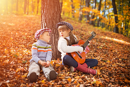 Kids sitting in autumn forest, playing guitar and singing. Kids are sitting by the tree, singing to each other. The sister is aged 7 and the brother is aged 3.