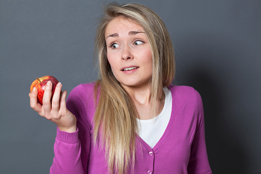 resigned young blonde woman with a red apple on her hands acting surprised
