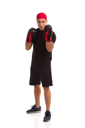 Confident man in sport shorts, black t-shirt and sneakers posing with boxing gloves. Full length studio shot isolated on white.