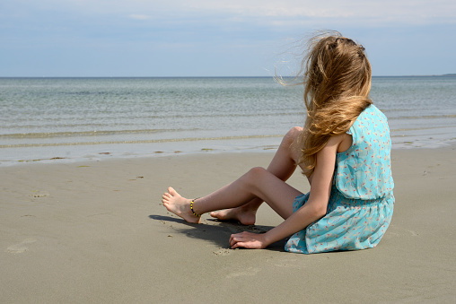 Side view of a teenage girl sitting barefoot on the sand at the sea. The girl has long blond hair and an northern european descent. She is wearing a light blue sundress. Shallow waves and calm sea under blue sky.