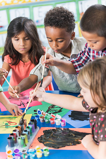 Group of preschoolers in art class painting pictures A group of preschool or kindergarten children in art class painting pictures at a table.  A little African American boy, 6 years old, is in the middle, reaching over to dip his brush in the paint. art class photos stock pictures, royalty-free photos & images