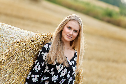 Beautiful young woman standing on field and leaning on roll bale of straw. She smiling and looking at camera. Shallow DOF. Shooting with the Canon EOS 5D Mark II.