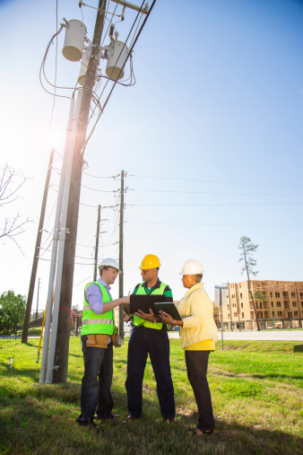 Utilities workers, engineer, and foreman talk at a job site.  Large apartments or office buildings under construction in background.