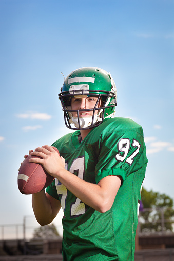 American football player quarterback in a game. A college American football player holding a football in his hand ready for throwing a pass to a receiver. He is wearing a green football jersey and helmet. Photographed in the stadium in vertical format with copy space available in the upper portion of the frame.