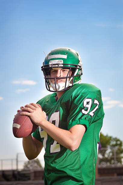 American Football Player Quarterback Throwing a Pass Close-up American football player quarterback in a game. A college American football player holding a football in his hand ready for throwing a pass to a receiver. He is wearing a green football jersey and helmet. Photographed in the stadium in vertical format with copy space available in the upper portion of the frame. american football ball photos stock pictures, royalty-free photos & images