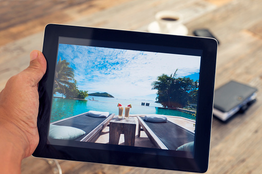 Man holding digital tablet with vacation photograph. He could be searching a travel website for hotels or resorts. Home interior setting with a wood textured table in the background. There is also coffee and a notebook in the background.