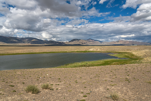 Scenic view of a beautiful lake in the desert with trees and vegetation on the banks on a background of mountains, blue sky and clouds