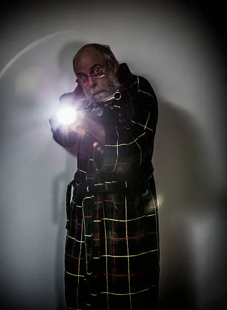 A trapped and terrified senior adult man wearing a plaid bathrobe is trying to aim a loaded shotgun at the camera while fumbling with a bright flashlight. Apparently he's caught the intruder or burgler criminal he'd been searching for in this dark basement room, but now he's cornered - suddenly realizing he's never fired a weapon before!