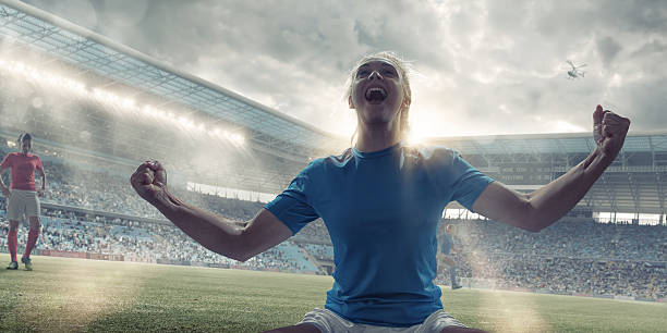 Womens Soccer Player Celebrating After Scoring A close up cross processed image of a professional woman soccer player kneeling on the grass with her arms out, fists clenched and head back, shouting in happiness and celebration at winning. The player is in a generic floodlit stadium full of spectators under a cloudy evening sky.  womens soccer stock pictures, royalty-free photos & images