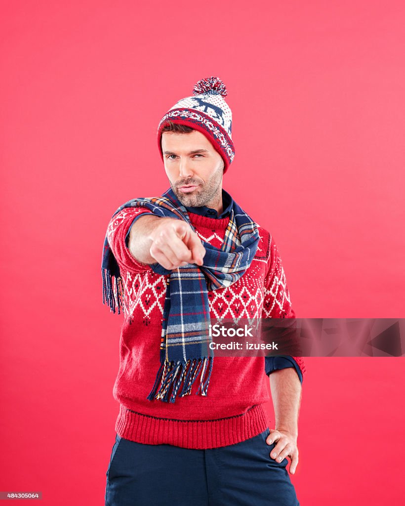 Happy man in winter outfit against red background Studio portrait of happy man in winter outfit - cap, scarf and sweater, standing against red background and pointing with index fingers at the camera. Men Stock Photo