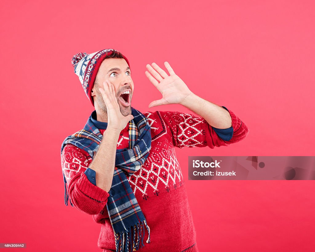 Happy man in winter outfit against red background Studio portrait of happy man in winter outfit - cap, scarf and sweater, standing against red background and screaming with raised hands. 2015 Stock Photo