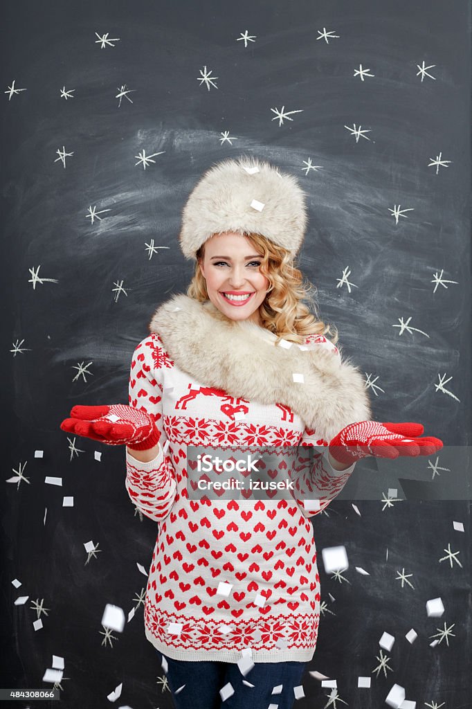 Happy blonde woman in winter outfit against blackboard Studio portrait of happy young woman in winter outfit - fur cap and scarf, sweater and gloves, standing against blackboard decorated with snowflakes, smiling at camera. 2015 Stock Photo