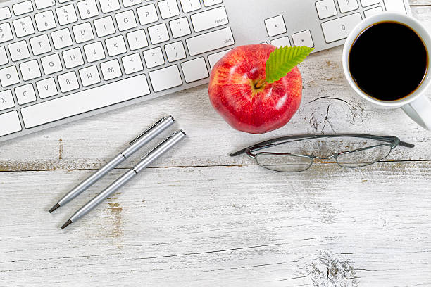 Red apple, coffee, reading glasses and silver pens next to computer keyboard with rustic white desktop underneath.