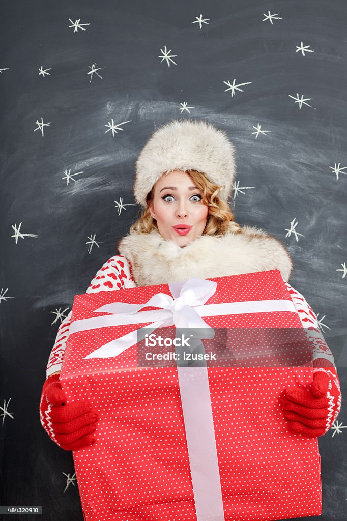 Happy blonde woman in winter outfit with christmas gift Studio portrait of surprised young woman in winter outfit - fur cap and scarf, sweater and gloves, standing against blackboard decorated with snowflakes and holding a big christmastgift, staring at camera.  Adult Stock Photo