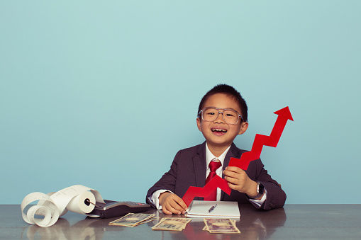 A young Japanese boy and businessman sits at his desk holding a line graph arrow showing his business is making money and growing. Boy dressed in business suit. Retro styled.