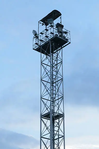 High lighting tower with observation platform at the top against a blue sky with clouds.