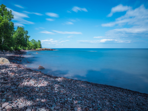 This is the shore of Lake Superior in Lutsen, Minnesota. This is a long exposure image leading to a blur or sense of motion of the clouds. The water also acquires a glassy appearance.