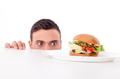Cheerful guy is hiding behind a table and peeping through it. He is looking at an unhealthy hamburger with desire. Isolated on background