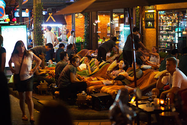 Bangkok, Thailand - March 11, 2015: Tourists are sitting outside of massage shop in second part of Khaosan Road at night and are getting foot massage. Thai people are doing massage. Other tourists are standing in street.
