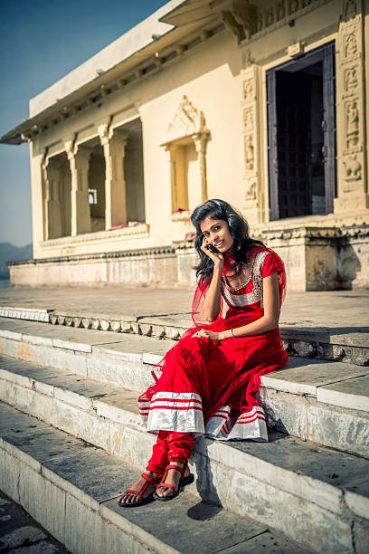 Young Indian woman sitting on Ambrai Ghat steps, Udaipur Woman in traditional dress enjoying morning at the temple on Pichola Lake, Ambrai Ghat in Udaipur, Rajasthan. Talking on her mobile phone. india indigenous culture indian culture women stock pictures, royalty-free photos & images