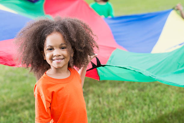 African American girl at summer camp holding parachute A cute little 5 year old African American girl at summer camp, having fun playing with a colorful parachute.  She is looking at the camera, smiling. 4 year old girl stock pictures, royalty-free photos & images