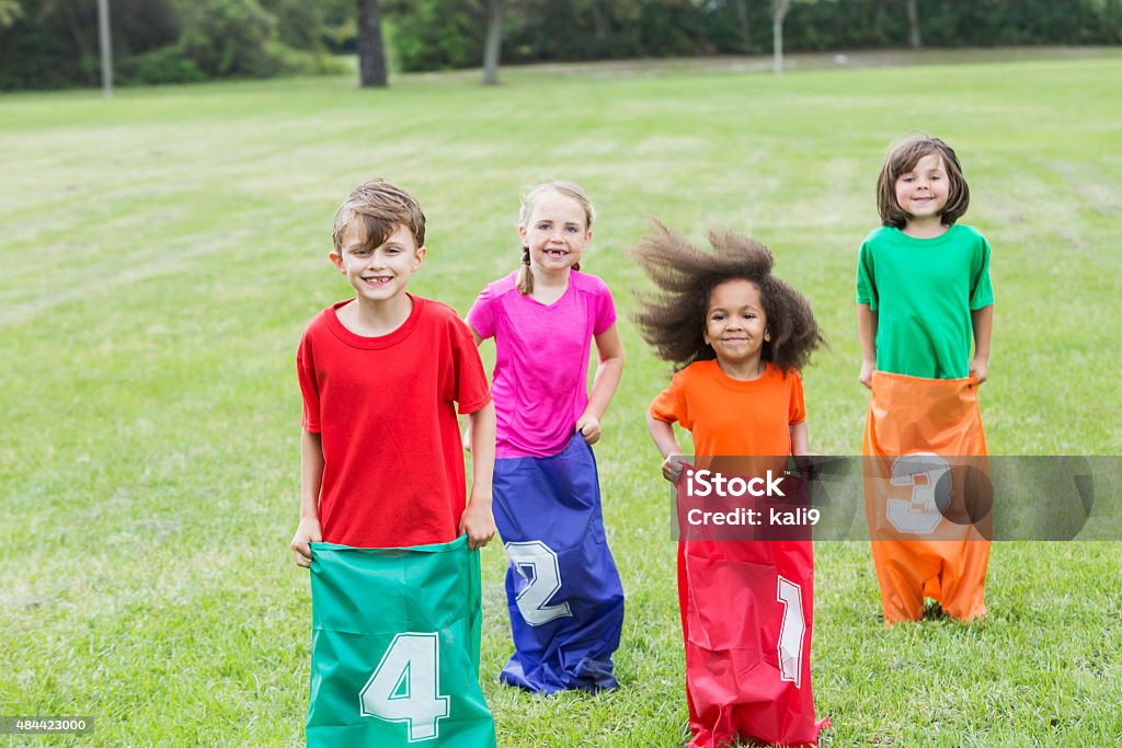 Four multiracial children in potato sack race A multiracial group of four children in a potato sack race in the park.  They are wearing colorful shirts and jumping in colorful, numbered sacks. Sack Race Stock Photo