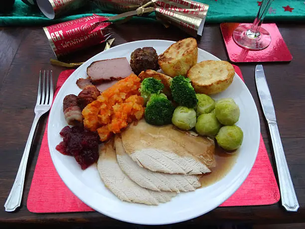 Photo showing a Christmas dinner place setting, with an attractive plate of food.  This traditional festive meal has been served on a white plate and includes roast turkey slices, roast potatoes, winter vegetables (Brussels sprouts, broccoli and a carrot-swede mash), stuffing, a chipolata sausage with bacon, and a spoon of cranberry sauce.
