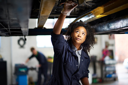 A young female adult mechanic is standing under a car in a garage. She is standing proudly and looking to camera