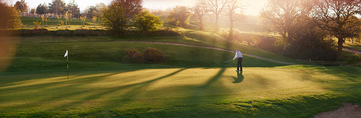 a lone golfer putts on the green , the low sun is coming from behind him and flaring to camera.