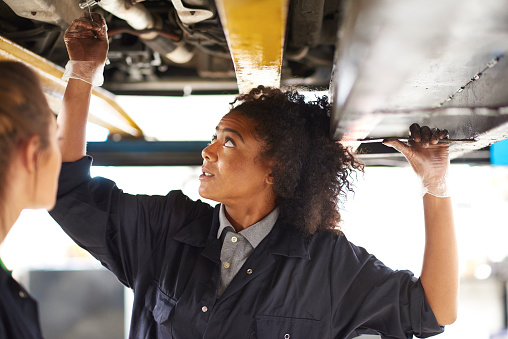 Two young female  mechanics are standing under a car on an inspection ramp in a garage. They are wearing blue overalls and holding a socket spanner . One mechanic is advising the other about the work they are doing .