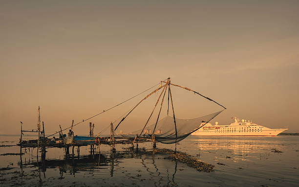 Fishermen, Chinese fishing net, and a cruise liner, Kerala, India. Fort Kochi, India - January 06, 2012: Fishermen watch a modern cruise lines enters the harbour as an ancient Chinese fishing net lays dormant at dusk on January 06, 2012, Fort Kochi, Kerala, India. cochin kerala india chinese fishing net stock pictures, royalty-free photos & images
