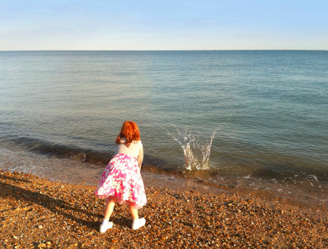 Image showing a young red headed girl, wearing a pink dress and grey cardigan, throwing pebbles into the sea on the main beachfront of Portsmouth, England, UK.
