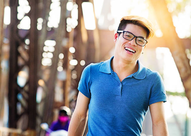 Young happy handsome Vietnamese man smiling while crossing Long Bien bridge in Hanoi, Vietnam. He is wearing a blue polo shirt, glasses and has hightlights in his hair. Back lit by the sunset.