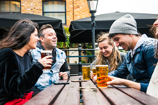 Group of friends hanging out in a London Pub seated on the outdoor tables. Their drinking a mix of blonde and red beers and talking together.