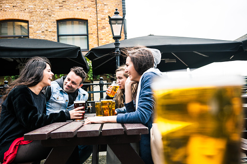 Group of friends hanging out in a London Pub seated on the outdoor tables. Their drinking a mix of blonde and red beers and talking together.