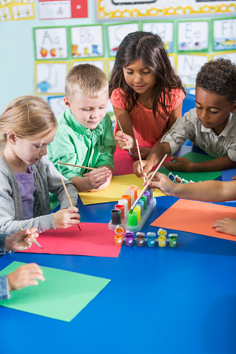 A multi-ethnic group of children in preschool or kindergarten class working on an art project.  The girls and boys are sitting at a table, reaching for paint with paintbrushes.
