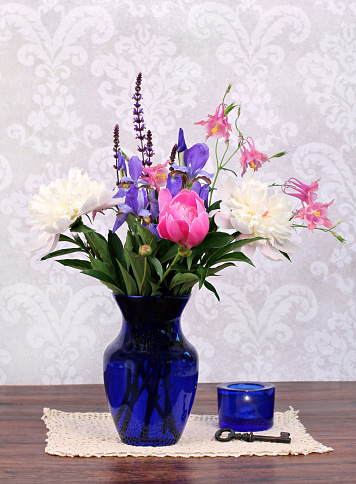 An assortment of typical and beautiful spring flowers in a navy vase with candle and antique key to the side. Arrangement on an antique doily.