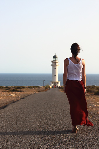 Young beautiful woman walking slowly to Cap de Barberia's lighthouse during an amazing summer sunset.