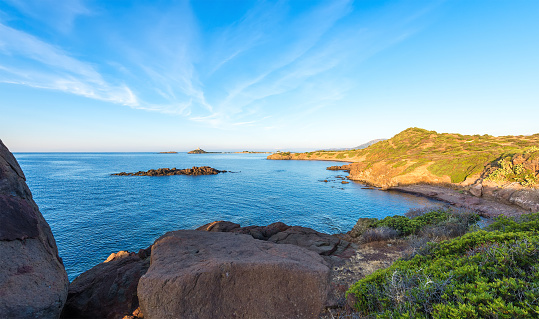 Panoramic view from cliffs to the sea, rocky coast and Nora tower near Pula in Sardegna, Italy. Wide angle wild nature shore landscape with dawn, green bush, stones and water cave.
