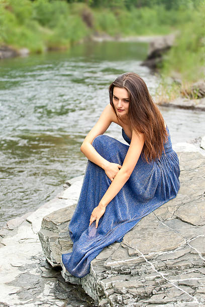 Pensive young attractive woman in long blue dress is sitting on a rock by the river. Blurred background of the river and green nature, she is looking down to river and thinking or dreaming. Summer atmosphere. Shooting with Canon EOS 5D Mark II.