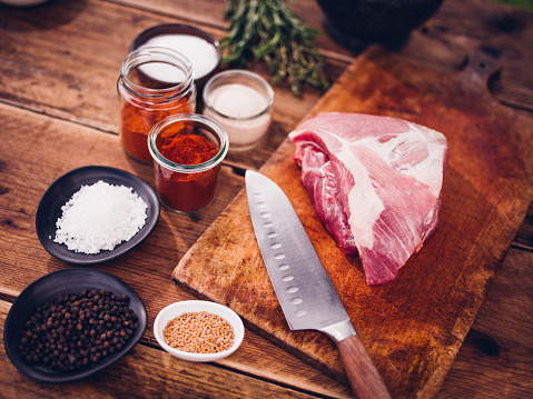 High angle view of a quality piece of raw pork on a wooden board with bowls of pepper, salt, paprika and herbs ready for seasoning on a vintage wooden table