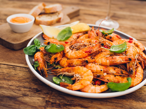 Spicy barbecued prawns with basil leaves and chillies presented on a plate with bread and sauce in the background on a vintage wooden table