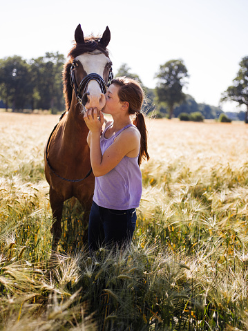 Teen girl lovingly giving her beautiful healthy horse a kiss on it's nose while it stands beside her peacefully in a wheat field