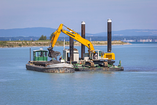 Bregenz, Austria - July 31, 2015: A Dredger is working to clear the River Rhine at the beginning of the Lake Constance (Bodensee) near Bregenz in Austria. A ship takes a scoop of sludge from the river, part of dredging activities. The flow of cold, mountain water with stones and sludge continues for some distance into the lake Bodensee. It is necessary to excavate this rubble.