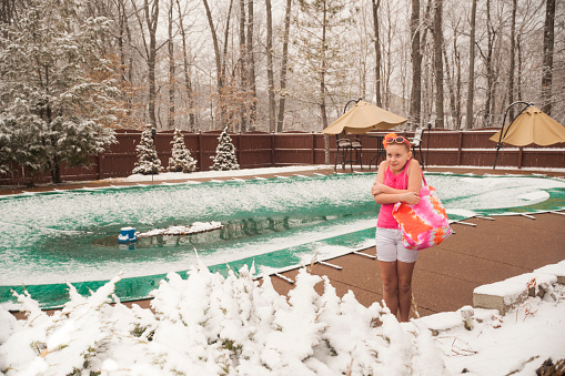 Preteen girl in summer short outfit stands in a snowy backyard next to a closed swimming pool during an unexpected snowfall in late March during a snowy cold spring break in southern Indiana