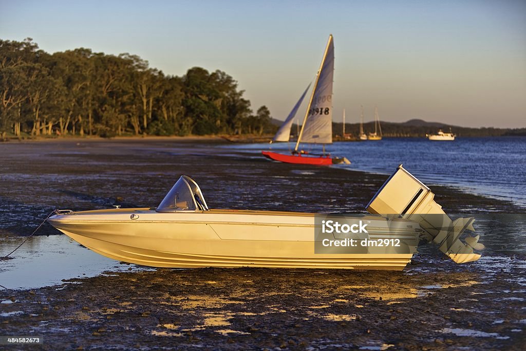 Speedboat on shore at low tide Moored speedboat which has become totally beached by the receding tide. Taken in golden late afternoon light. Sailboat in background. Day Stock Photo