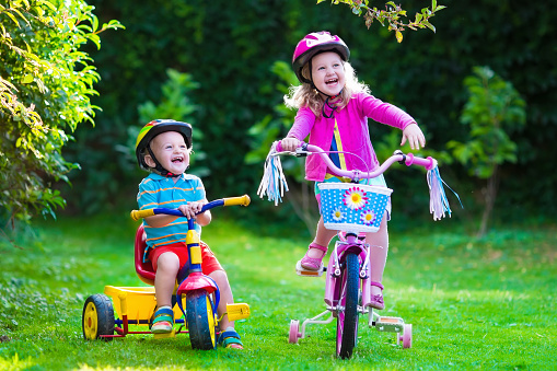 Kids riding bikes in a park. Children enjoy bike ride in the garden. Girl on a bicycle and little boy on a tricycle in safety helmet playing together outdoors. Preschool child and toddler kid biking.