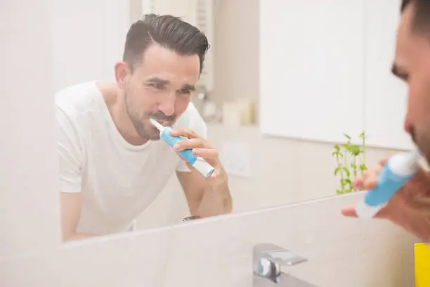Handsome man brushing teeth and smiling to the mirror. He is wearing white t-shirt. He is latino with a black beard.