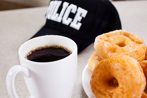 Policeman has taken a morning break from his patrol to eat donuts and drink coffee at a local restaurant.  His policeman hat is on the cafe countertop. No people in image. Law enforcement. Breakfast. 
