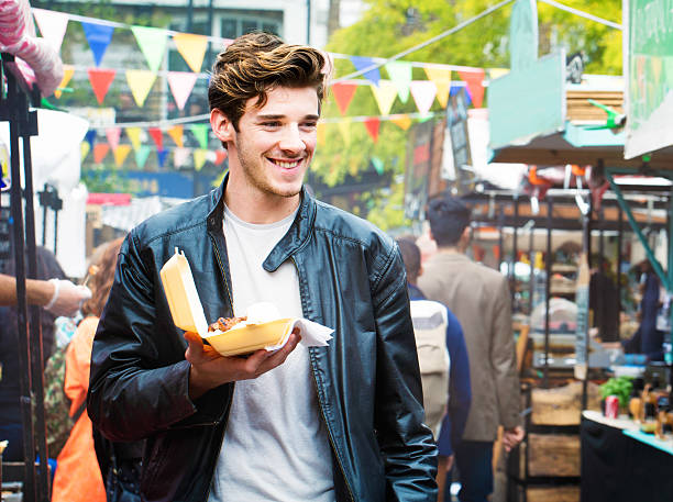 Handsome British man walks through fair market with street food Handsome British man walks through fair market with street food in hand and a big smile. He is wearing a leather jacket an long wavy hair. Photographed in London, UK. english cuisine stock pictures, royalty-free photos & images